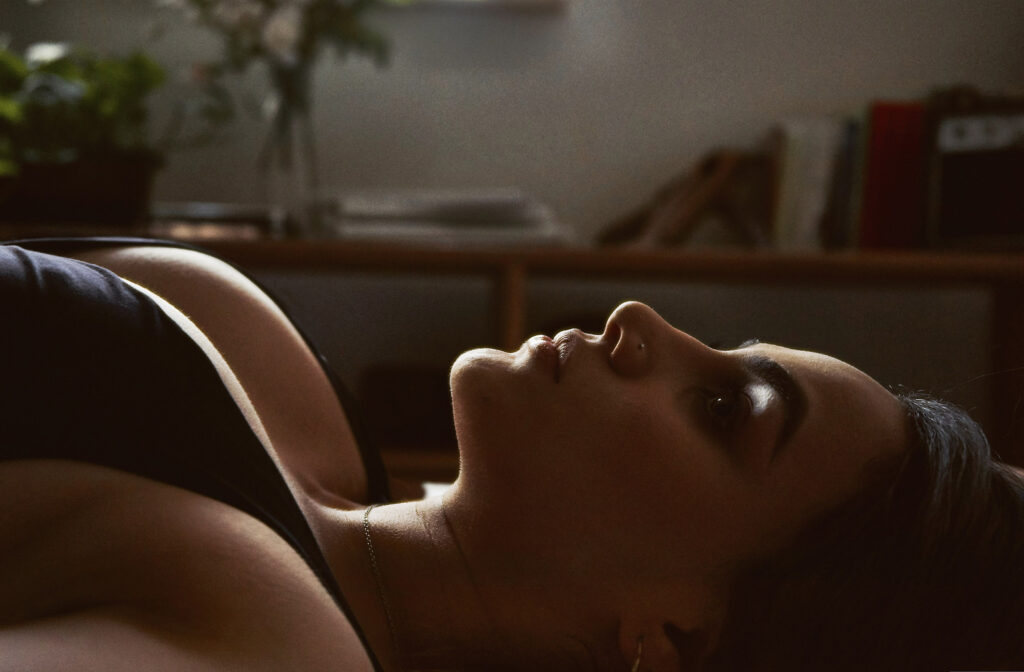 Valeria Cisneros lies on her side gazing upwards with a serene expression. The room is dimly lit with sunlight filtering through a window casting a soft glow on her face. A table with books and a vase of flowers is visible in the background.